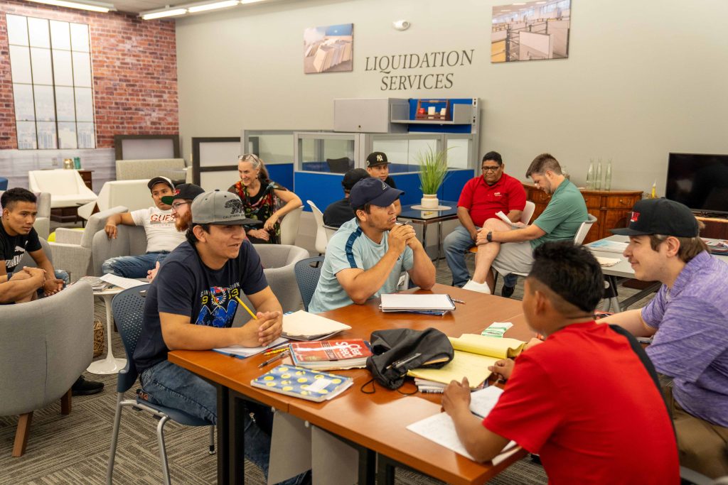 Group of Turnkey Workplace Services employees gather in the company's showroom four nights a week to learn English. Employees are seated at tables and in armchairs, with notebooks, speaking English to each other.