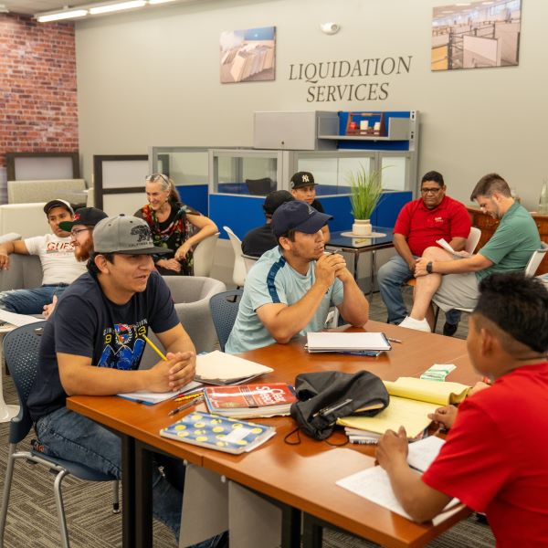 Group of Turnkey Workplace Services employees gather in the company's showroom four nights a week to learn English. Employees are seated at tables and in armchairs, with notebooks, speaking English to each other.