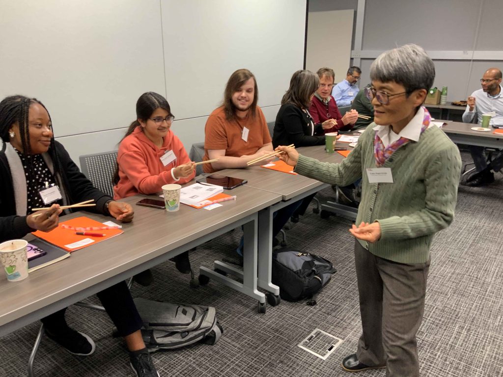 Teacher demonstrates how to hold and use chopsticks with group of students.