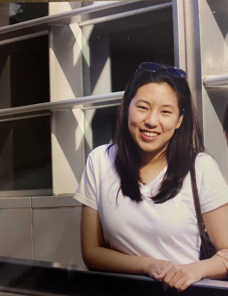 Young Asian woman leaning on railing with sun behind her.